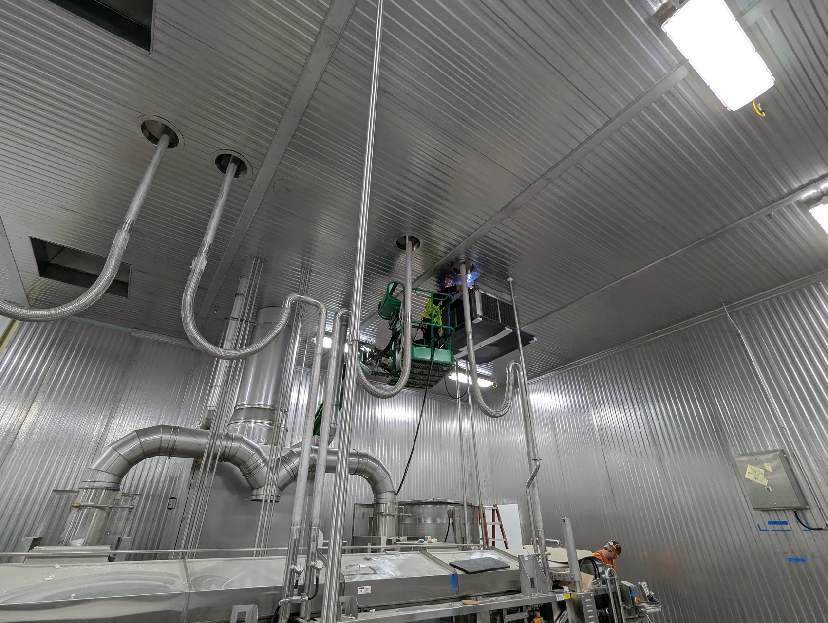 Polished stainless spiral ductwork runs across a food processing ceiling with a welder on a lift overhead